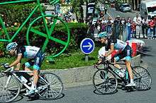 A pair of road racing cyclists in the team's black, white, and blue jersey, riding around a traffic island that features a large green sculpture of a bicycle. Spectators and motor vehicles are visible behind roadside barricades in the background.