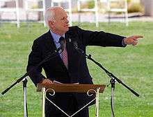 White-haired man standing at podium and speaking and gesturing with outstretched arm and an outdoor venue