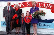 White-haired man, elderly white-haired woman, young boy, young girl, short-haired woman holding roses, all in front of sign showing a ship's silhouette