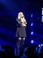 A young blonde woman singing into a microphone onstage, sporting a black skirt and black Bad Gal jacket. Blue stage lights shine upon her, while the logo of iHeart Radio serves as her backdrop.