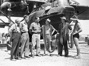 Three military personnel in slouch hats with lines of machine-gun belts wrapped around them speak with three civilians in front of a twin-engined aircraft
