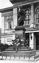 black-and-white photograph of a statue of a robed male figure on a stepped pedestal, inscribed 'Felix Mendlessohn Bartholdy', with a seated female figure holding a lyre at its base, in front of an arcaded building
