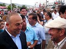 Two older Turkish men stand facing each other, one bald, the other wearing a white cap, while a large crowd mingles behind them along a waterfront.