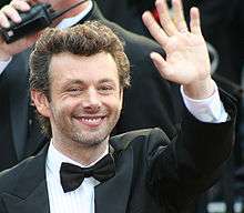 Michael Sheen, a caucasian male in his early-40s with dark curly hair, wears a black suit and white shirt with a black bow-tie. He smiles and waves with his left hand.