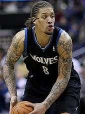 Michael Beasley with cornrows, wearing a black Timberwolves uniform with blue trim and griping a basketball with both hands in the lower left