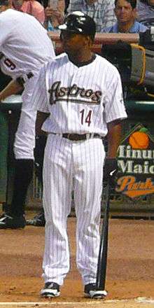 A dark-skinned man in a white pinstriped baseball uniform stands on the dirt. He holds a black baseball bat in his left hand, and his jersey reads "Astros" in script lettering and "14" in block print.