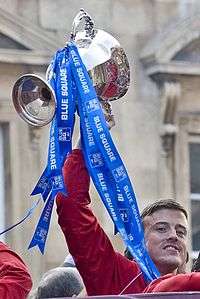 A man with dark hair is wearing a red jacket. He is holding aloft a trophy.