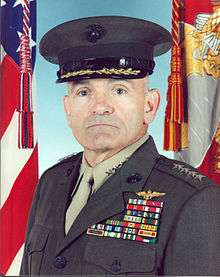 A color image of Michael Williams, a white male in his Marine Corps Service A uniform. He is wearing a hat, several ribbons are visible as well as naval aviator insignia. The Marine Corps flag and United States flag are visible in the background.