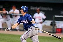 Michael Young, wearing a blue batting helmet and baseball jersey with the lettering TEXAS across it and the Flag of Texas on the left sleeve, clutches a baseball bat in his left hand and looks towards the left after hitting a baseball.