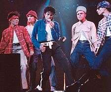 Five males standing next to each other. The male to the farthest left is wearing a red hand band and a red checkered shirt with grey pants. The male to the second left is wearing a red hat and a white shirt. The male in the center is wearing a blue jacket and black pants with a white belt. The male to the second right is wearing a white hat and a white shirt and blue pants. The male to the farthest right is wearing a blue bandana and blue pants.