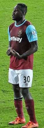 A black man wearing a claret and blue football shirt, white shorts, claret socks and orange football boots stares ahead, his hands held together in front.