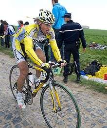 A cyclist wearing a yellow and gray cycling jersey, riding over cobblestones. Spectators on the roadside are looking behind him, out of the frame.