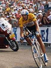 A man on a bicycle wearing a helmet with a visor and a yellow top with black shorts. In the background there is a red motorcycle with two men on it.
