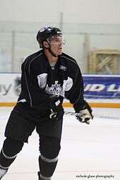 A hockey player in a black and white uniform with a crown logo on his chest looks to his left as he skates.