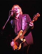 Mike Mills plays bass guitar and sings into a microphone while wearing a Nudie suit