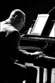 A three-quarters view of Mike Garson—a Caucasian male with a shaved head wearing a suit and sunglasses—seated at a piano