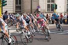 A large group of cyclists makes a right turn around a street corner in a major city. Prominent among them is a cyclist wearing an all-pink jersey, which represents being the overall leader in the race. A crowd of spectators watches the group of cyclists from the roadside.