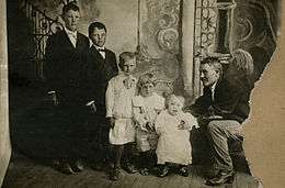 Five children standing in descending order of age and height, adult male sitting next to the youngest one