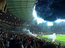 A pitch level perspective of a floodlit oval stadium, filled with people wearing black and white clothes and raising their arms; smoke from flares fills the air, and a section of the playing field and a goal can be seen.