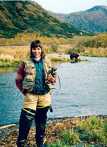 Woman holding binoculars, in thigh waders, by stream with back to bear