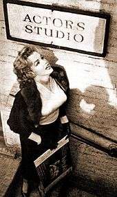 Monroe, who is wearing a skirt, blouse and jacket, standing below a sign for the Actors Studio looking up towards it