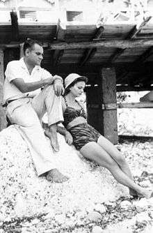 Elsa Morante is leaning against a large rock with Alberto Moravia sitting on the rock, at the beach in Capri, 1940s
