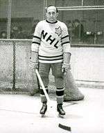 An ice hockey player stands on an ice rink. He is wearing similar equipment as before, except that his sweater has the letters "NHL" in a downward diagonal with a large star on his left shoulder.
