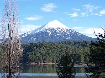 A conical, snow-capped mountain rises above a forest and a lake.