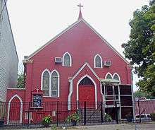 A one-and-a-half-story red wooden church with pointed roof seen from the front. There is a cross on top and a black iron fence in the front.