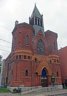 An ornate red brick church with rusty orange trim. A central steeple rises from between two octagonal towers