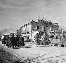 Soldiers march along a road past a stone building that has been damaged
