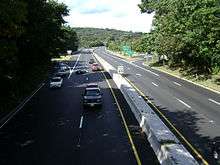 An overhead view of a four-lane divided freeway running through a wooded area. An interchange is seen in the distance.