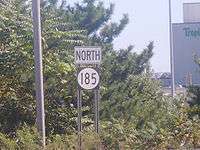 A road sign reading north above a black rectangle with a white oval containing 185 in black numerals. Trees and industrial buildings can be seen in the background.