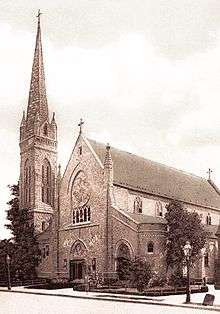 A sepia-toned photograph of a church's stone facade in the Gothic style. Centered and above the doorway is a large, circular, stained glass window. Attached to the left side of the building is a bell tower spire.