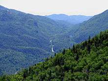 A narrow two-lane highway runs along the base of a valley bounded on each side by many large, tree-covered mountains.