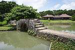 A pond crossed by a stone causeway with a bridge. There is a low wooden house beyond the pond.