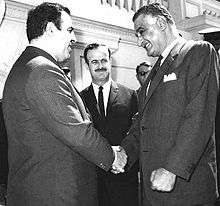 Two men shaking hands, with mustachioed man in background