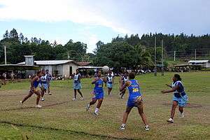An outdoor netball game taking place in Fiji. The game is being played on grass. The players are either high school age or adults. Both teams are wearing blue netball uniforms. The team in the darker blue is in the act of passing the ball.