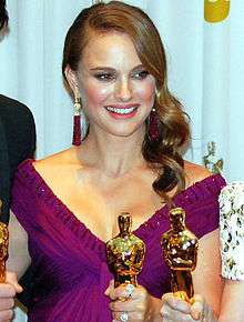 A young woman with brown hair dangling to her left in a V-neck purple dress holding one of the golden statuettes seen above