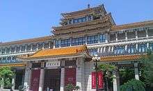 An ornate building front stretching the width of the image, slightly tilted, against a uniformly blue sky. Its front has a projecting pavilion in the Chinese style, echoed by a similar pagoda-style top on the roof above it.