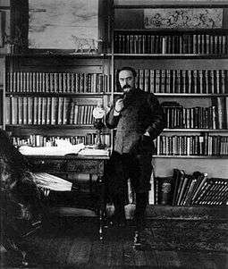 Full length photo of a man dressed in a khaki jacket standing in a book lined study. He has short hair, a moustache and is smoking a pipe.