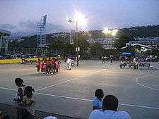 Two netball teams huddled together on an outside netball court. The court is lit by lights as it is twilight.