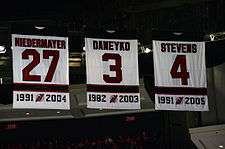 Three banners with the New Jersey Devils retired numbers, showing the player surname and years with the team: Niedermayer, 27, 1991–2004; Daneyko, 3, 1982–2003; and Stevens, 4, 1981–2005.
