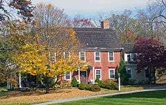 A brick house with rounded black roof and two chimneys behind two trees, one of which has lost half of its yellowed leaves which blanket the ground below, the other of which is dark red. The house has a wooden wing on the right and a less visible covered porch on the left