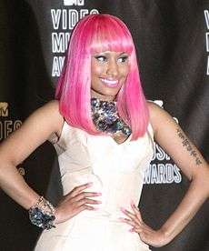 A tan-skinned woman in a bright pink wig poses with hands on both side of her hips. Smiling, she stands before a black background and has Mandarin characters tattooed on her right arm.