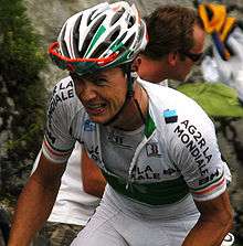 A road racing cyclist wearing a mostly white jersey with black trim and a green clover on the midsection. He has a pained grimace on his face, and a spectator is visible in the background.