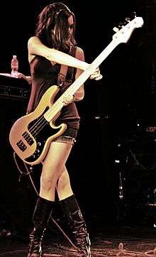 Nicole Fiorentino—a brunette Caucasian woman with long hair—plays bass guitar onstage and is wearing a dress, fishnet stockings, and boots