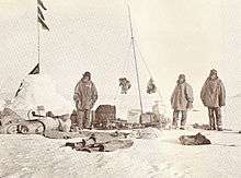  On the left is a snow cairn with flags. Three men are nearby, and assorted equipment is strewn on the snow.