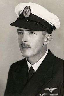 Head-and-shoulders portrait of man with moustache wearing dark-coloured jacket with black-and-white peaked cap