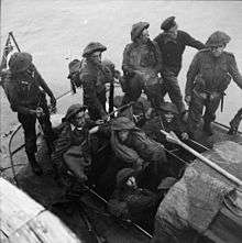 A black and white photograph showing soldiers wearing combat equipment sitting in a small watercraft beside a pier